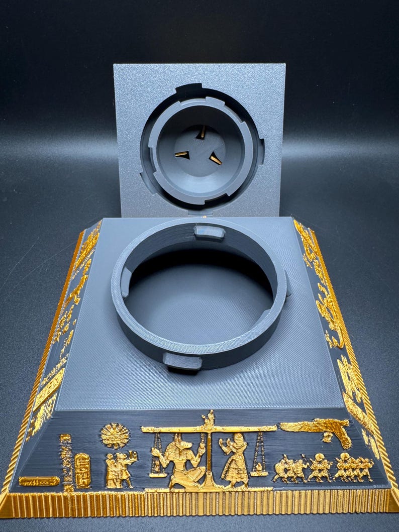 Puede incluir: Un soporte gris impreso en 3D con una abertura circular y una tapa separada. El soporte presenta dise&ntilde;os jerogl&iacute;ficos egipcios dorados a lo largo de los bordes y la base, con un relieve detallado de figuras y s&iacute;mbolos.