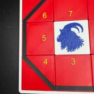 May include: A red game board with black trim and a blue goat silhouette in the center. The numbers 3, 4, 5, 6, and 7 are printed in yellow on the board.