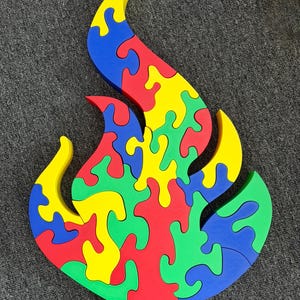 Puede incluir: Un rompecabezas de madera multicolor con forma de llama, que presenta piezas entrelazadas en tonos vibrantes de rojo, amarillo, verde y azul. El rompecabezas est&aacute; construido con segmentos gruesos de bordes lisos que encajan para formar un dise&ntilde;o de fuego.