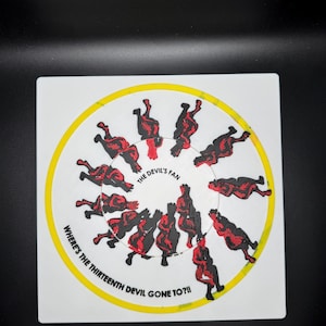 Op de afbeelding: Vierkante witte onderzetter met een gele ring en de tekst "WHERE'S THE THIRTEENTH DEVIL GONE TO?!!" en "THE DEVIL'S FAN". De onderzetter heeft een cirkelvormig ontwerp van rode en zwarte duivelsfiguren.