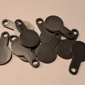 May include: A group of small, dark grey, paddle-shaped items. Each has a circular head with a ridged texture and a handle with a hole. The objects are scattered on a light background. The items appear to be made of plastic.
