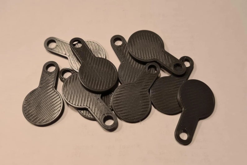 May include: A group of small, dark grey, paddle-shaped items. Each has a circular head with a ridged texture and a handle with a hole. The objects are scattered on a light background. The items appear to be made of plastic.