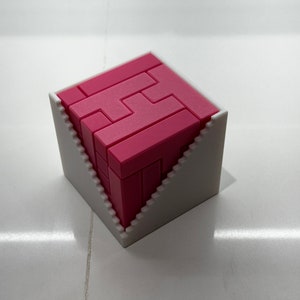 Puede incluir: Un rompecabezas 3D rosa en forma de cubo, ensamblado dentro de un marco blanco de forma triangular.