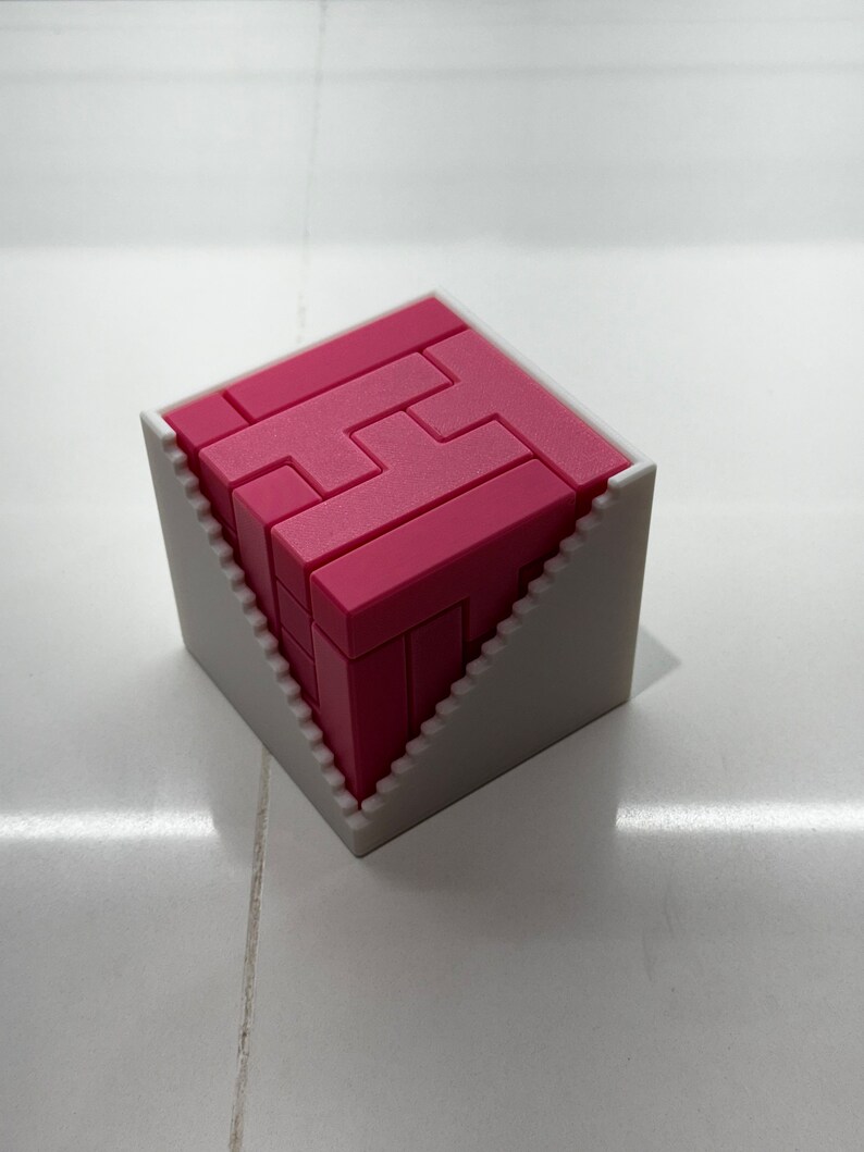 Puede incluir: Un rompecabezas 3D rosa en forma de cubo, ensamblado dentro de un marco blanco de forma triangular.
