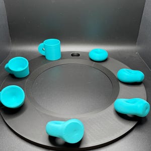 Op de afbeelding: Een verzameling turquoise 3D-geprinte objecten, waaronder kleine mokken en ringvormige items, gerangschikt op een zwarte cirkelvormige basis. De objecten zijn gelijkmatig verdeeld rond het midden, wat een visueel evenwichtige compositie cre&euml;ert. De achtergrond is effen zwart.
