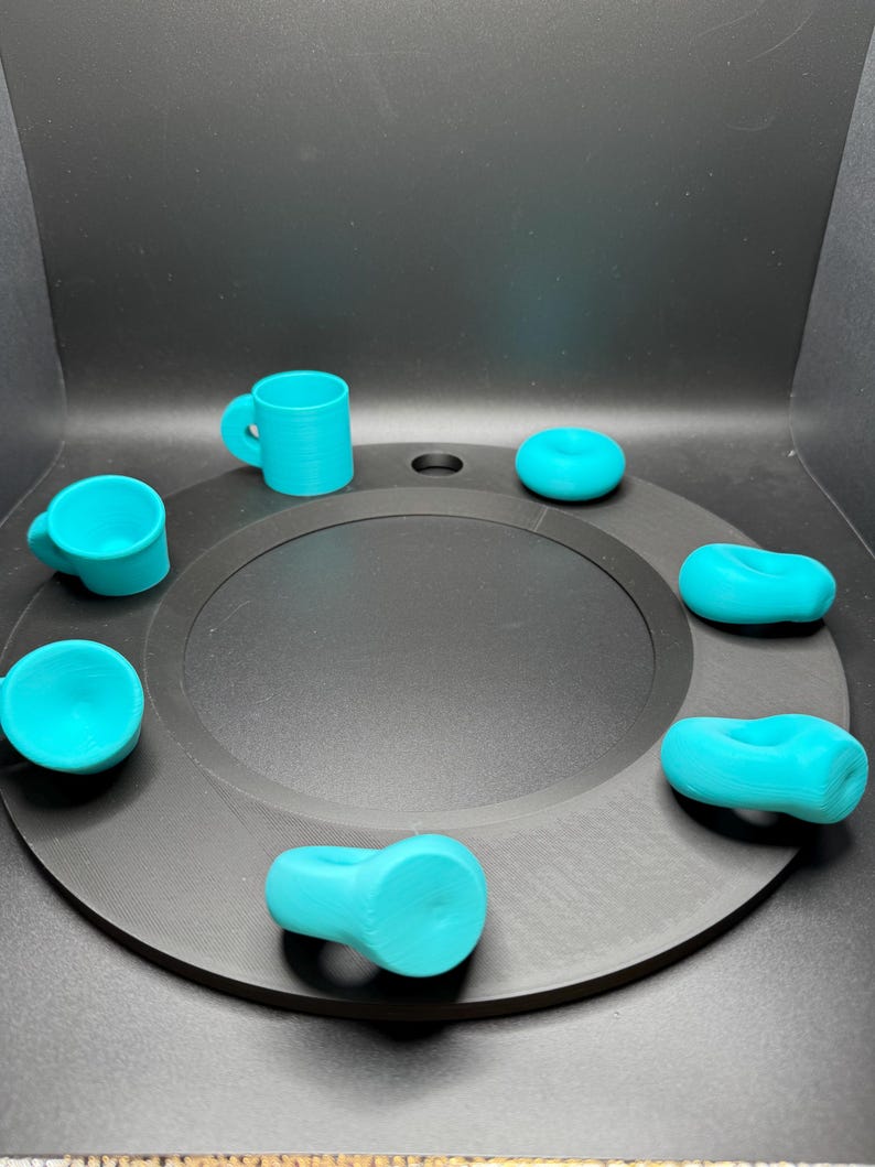 Op de afbeelding: Een verzameling turquoise 3D-geprinte objecten, waaronder kleine mokken en ringvormige items, gerangschikt op een zwarte cirkelvormige basis. De objecten zijn gelijkmatig verdeeld rond het midden, wat een visueel evenwichtige compositie cre&euml;ert. De achtergrond is effen zwart.