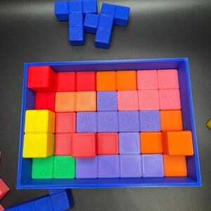 May include: A blue rectangular tray filled with colorful square blocks in shades of red, orange, pink, purple, green, and yellow. Two blue block shapes are positioned above the tray. This image showcases a puzzle game.