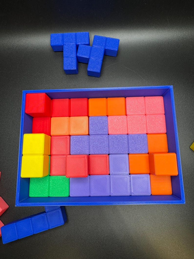 May include: A blue rectangular tray filled with colorful square blocks in shades of red, orange, pink, purple, green, and yellow. Two blue block shapes are positioned above the tray. This image showcases a puzzle game.