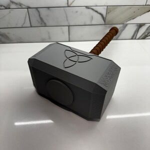Peut inclure: Une r&eacute;plique en plastique gris du marteau de Thor, Mjolnir, avec un manche en bois brun. Le marteau pr&eacute;sente un motif circulaire avec un symbole &agrave; trois pointes sur le dessus et des d&eacute;tails complexes sur les c&ocirc;t&eacute;s.