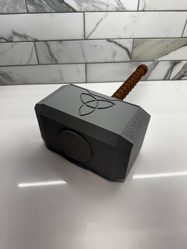 Peut inclure: Une r&eacute;plique en plastique gris du marteau de Thor, Mjolnir, avec un manche en bois brun. Le marteau pr&eacute;sente un motif circulaire avec un symbole &agrave; trois pointes sur le dessus et des d&eacute;tails complexes sur les c&ocirc;t&eacute;s.