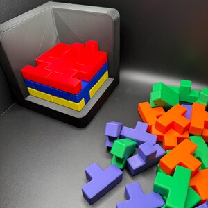 May include: A set of colorful plastic puzzle pieces in red, orange, green, blue, and purple. The pieces are shaped like the letter T and are arranged in a pile on a black surface. Some of the pieces are stacked in a small black box.
