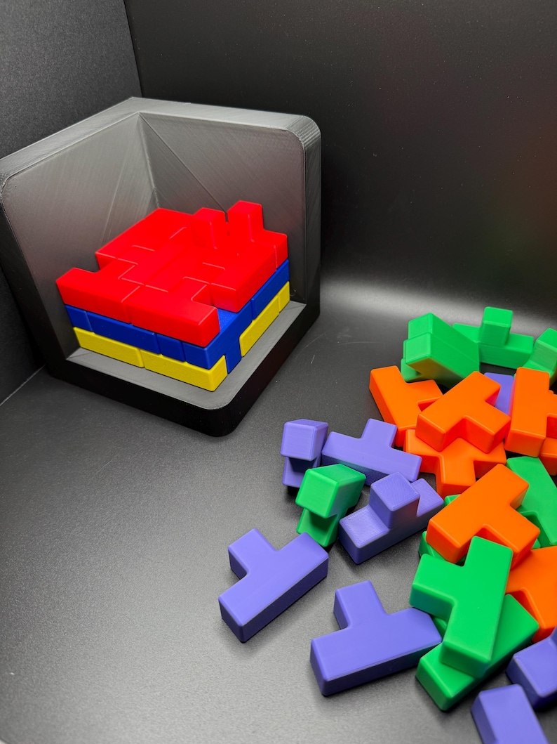 May include: A set of colorful plastic puzzle pieces in red, orange, green, blue, and purple. The pieces are shaped like the letter T and are arranged in a pile on a black surface. Some of the pieces are stacked in a small black box.