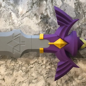 May include: A purple and gold plastic sword with a triangular design on the blade and a winged hilt. The sword is on a gray and white textured surface.