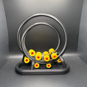 May include: A black, 3D-printed kinetic sculpture featuring two concentric circular tracks. Yellow and red cylindrical pieces are scattered within the tracks and base. The sculpture is designed to allow the pieces to move freely.