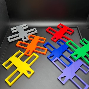 K&ouml;nnte beinhalten: Eine Sammlung farbenfroher, kreuzf&ouml;rmiger Objekte in verschiedenen Farben, darunter Gelb, Orange, Rot, Blau, Gr&uuml;n, Lila und Grau. Jedes Objekt hat einen Ausschnitt in der Mitte. Die Objekte sind auf einer dunklen Oberfl&auml;che angeordnet.