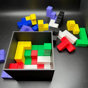 May include: A black square container filled with colorful, geometric puzzle pieces. The pieces are various shapes and colors, including red, white, green, yellow, blue, and purple. Some pieces are scattered outside the container.