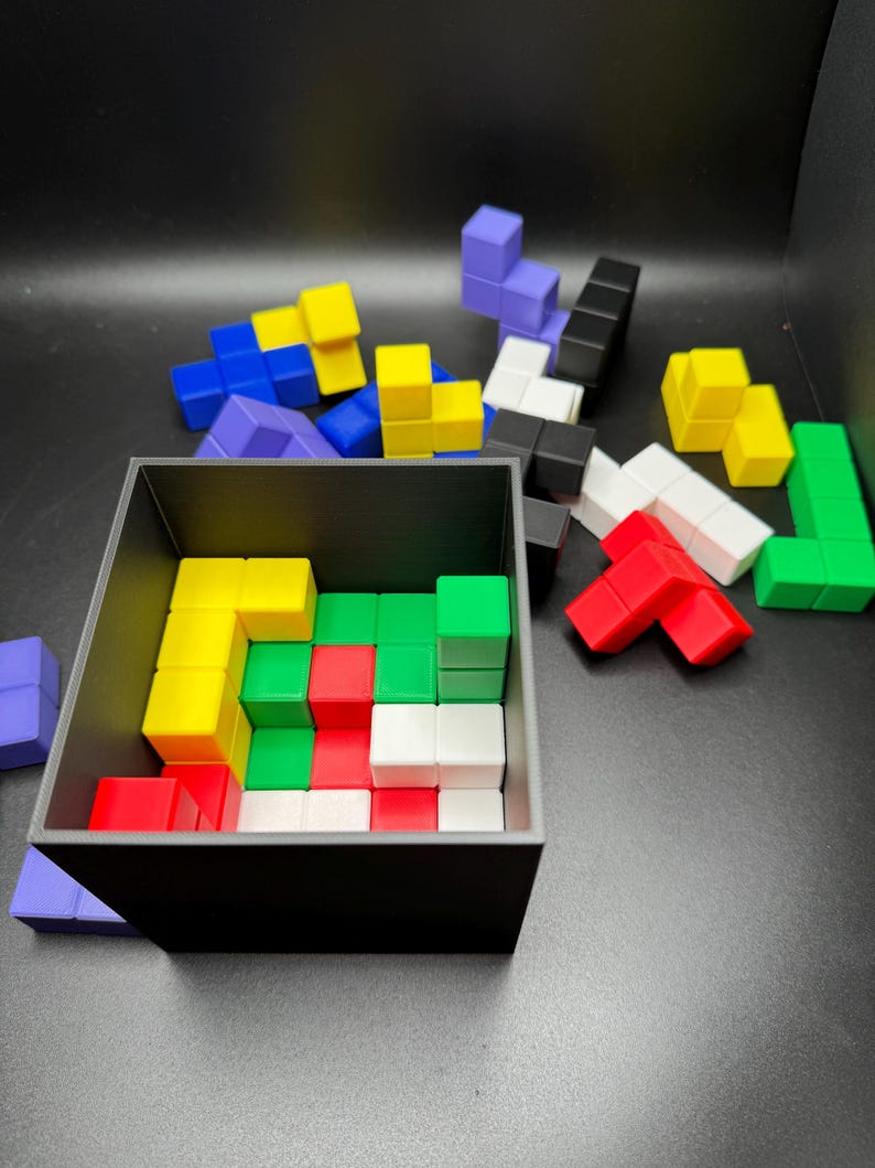 May include: A black square container filled with colorful, geometric puzzle pieces. The pieces are various shapes and colors, including red, white, green, yellow, blue, and purple. Some pieces are scattered outside the container.