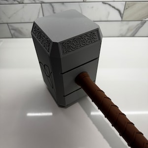 Peut inclure: Une r&eacute;plique en plastique gris du marteau de Thor, Mjolnir, avec un manche en cuir brun. La t&ecirc;te du marteau pr&eacute;sente des sculptures complexes et un motif en relief.