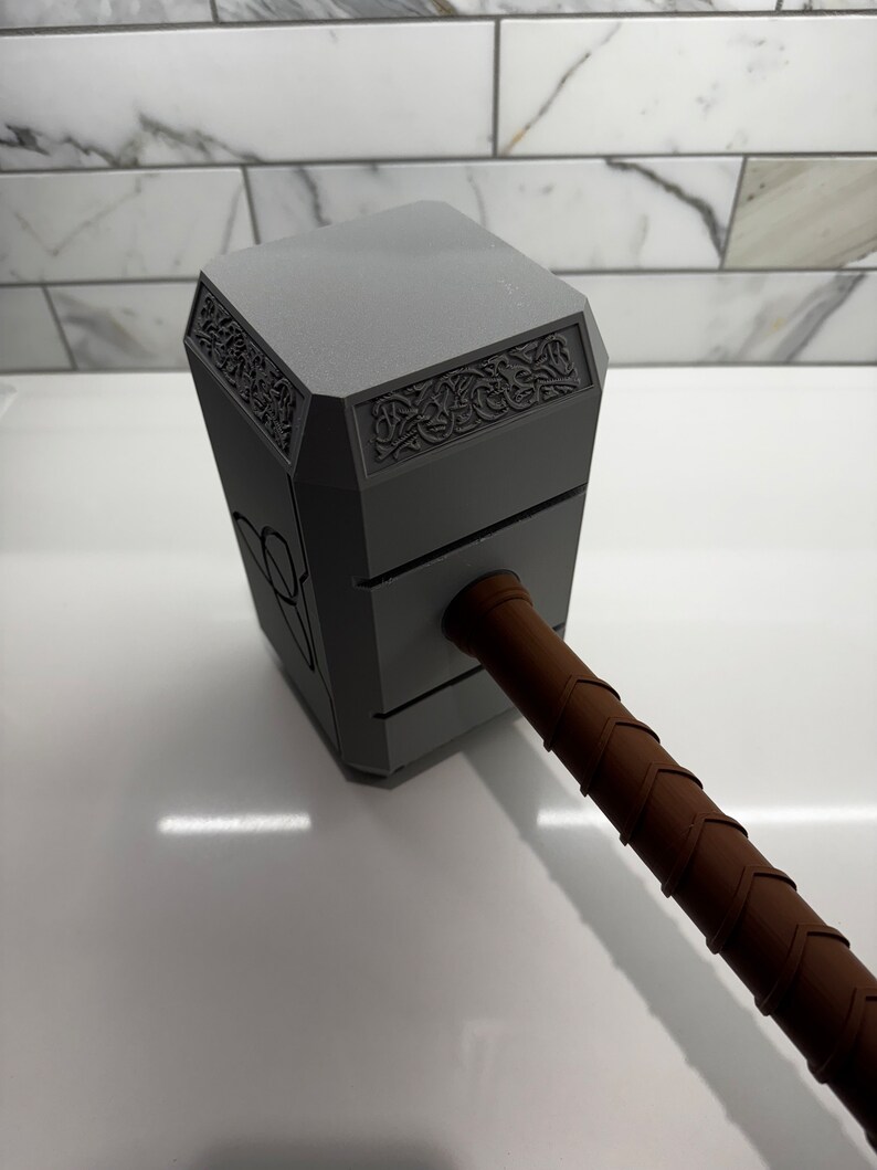 Peut inclure: Une r&eacute;plique en plastique gris du marteau de Thor, Mjolnir, avec un manche en cuir brun. La t&ecirc;te du marteau pr&eacute;sente des sculptures complexes et un motif en relief.