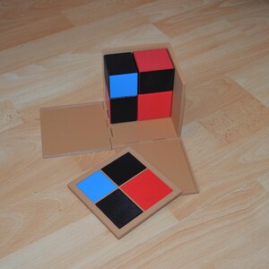 May include: A wooden box with a lid that opens to reveal a set of four colored blocks. The blocks are arranged in a square pattern, with two red blocks, one blue block, and one black block. The box is open and the lid is lying on the table next to the box.