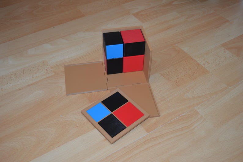 May include: A wooden box with a lid that opens to reveal a set of four colored blocks. The blocks are arranged in a square pattern, with two red blocks, one blue block, and one black block. The box is open and the lid is lying on the table next to the box.