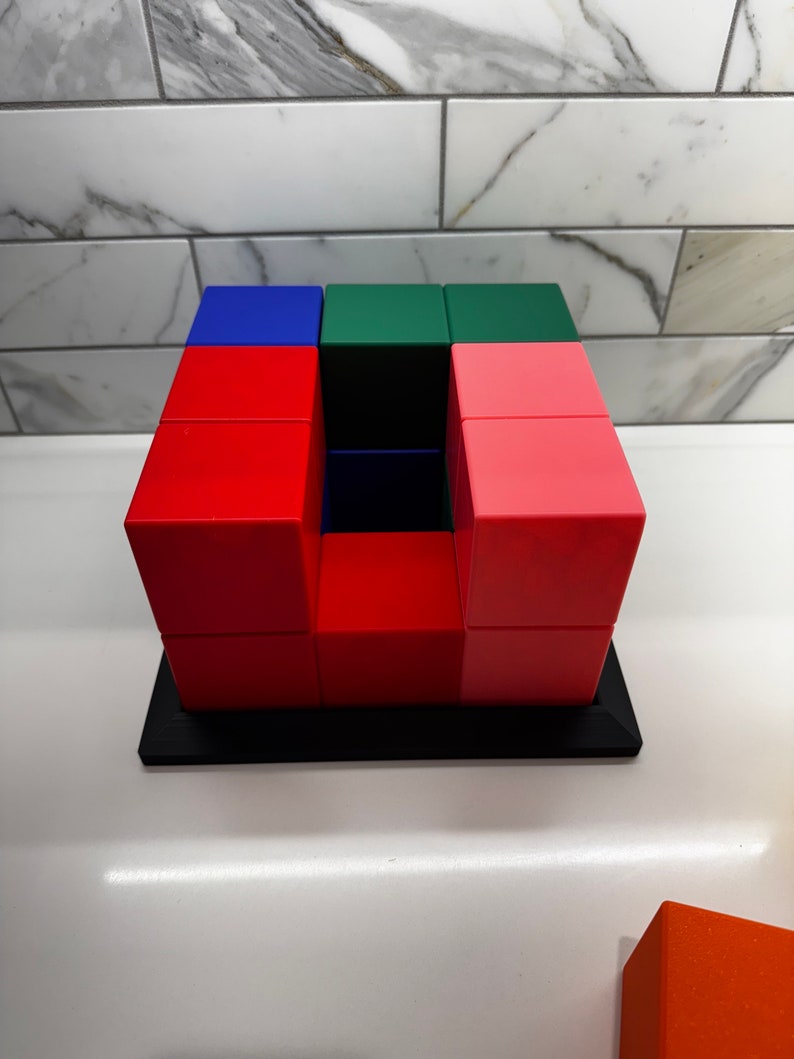 May include: A red, green, and blue cube puzzle with a hollow center. The puzzle is made of 12 individual cubes and is sitting on a black base.
