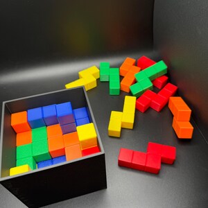 May include: A black box filled with colorful wooden blocks. The blocks are arranged in a Tetris-like pattern. There are several loose blocks outside the box, also in various colors.