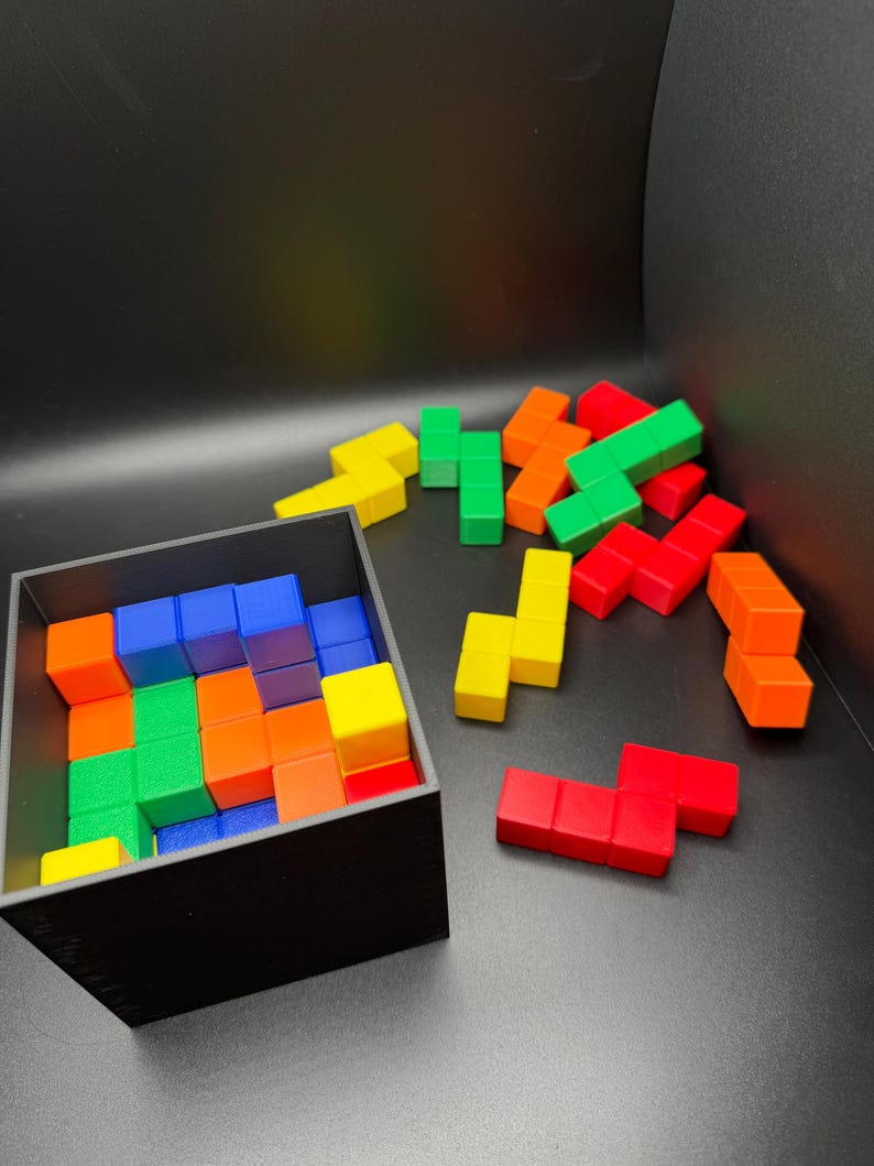 May include: A black box filled with colorful wooden blocks. The blocks are arranged in a Tetris-like pattern. There are several loose blocks outside the box, also in various colors.