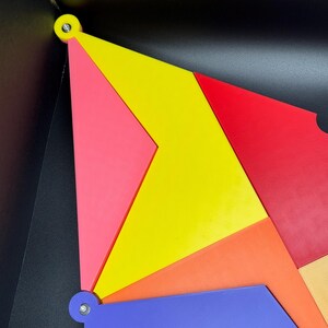 Peut inclure: Composition g&eacute;om&eacute;trique abstraite avec des formes vives et imbriqu&eacute;es en rouge, jaune, rose, orange et bleu. Les pi&egrave;ces sont fix&eacute;es avec de petites attaches argent&eacute;es sur un fond noir. Le design est moderne et color&eacute;.