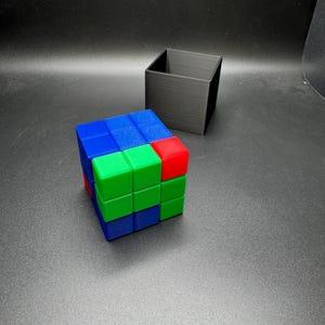 May include: A colorful Rubik's Cube with blue, green, and red sides sits next to a black square container. The cube is partially solved, showcasing the vibrant colors of the puzzle. The black container is empty.