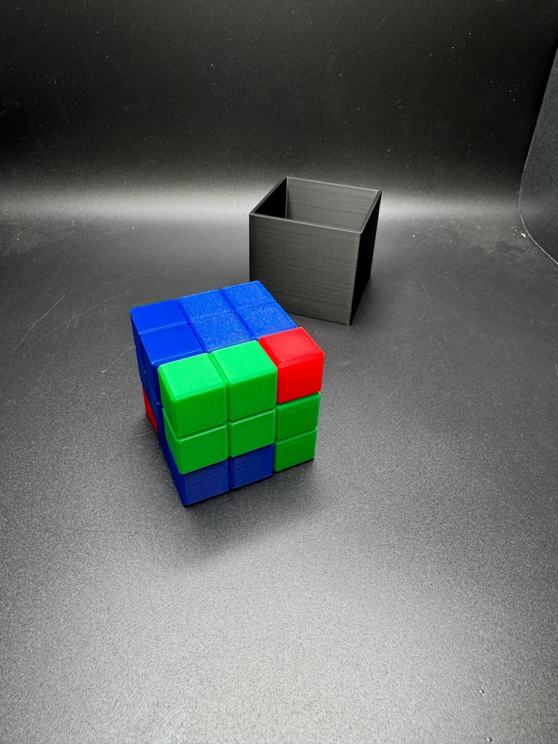 May include: A colorful Rubik's Cube with blue, green, and red sides sits next to a black square container. The cube is partially solved, showcasing the vibrant colors of the puzzle. The black container is empty.