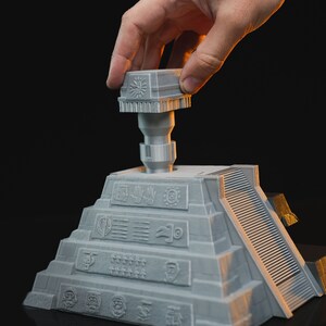 May include: A gray 3D printed model of a pyramid with a removable top piece. The pyramid has a series of symbols and designs on its sides. The top piece is a square with a symbol on it.
