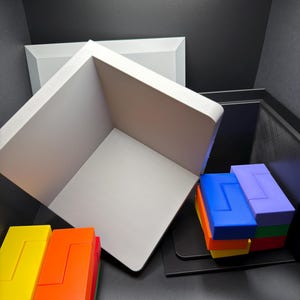 May include: A white open-faced cube structure positioned diagonally, accompanied by two stacks of interlocking rectangular blocks in vibrant colors including yellow, orange, red, green, blue, and purple, arranged on a dark surface.