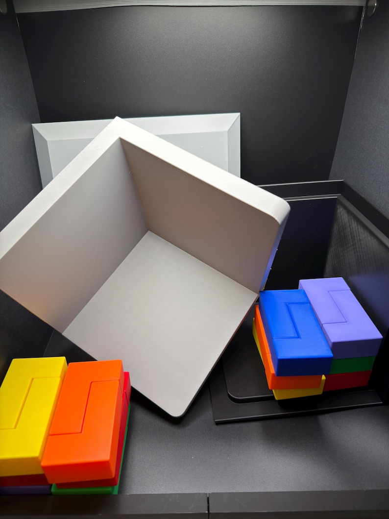 May include: A white open-faced cube structure positioned diagonally, accompanied by two stacks of interlocking rectangular blocks in vibrant colors including yellow, orange, red, green, blue, and purple, arranged on a dark surface.