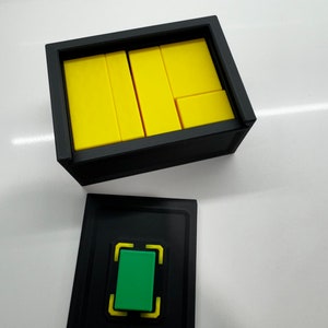 Pu&ograve; includere: Una scatola nera con il coperchio aperto, che rivela blocchi rettangolari gialli all'interno. La scatola ha una scatola nera pi&ugrave; piccola attaccata al fondo con un pulsante verde all'interno di una cornice gialla.