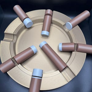 K&ouml;nnte beinhalten: Ein goldfarbener Aschenbecher mit kreisf&ouml;rmigem Design, der mehrere braune und graue zylindrische Objekte aufweist. Die Objekte sind um den Aschenbecher angeordnet. Der Aschenbecher hat eine strukturierte Oberfl&auml;che.