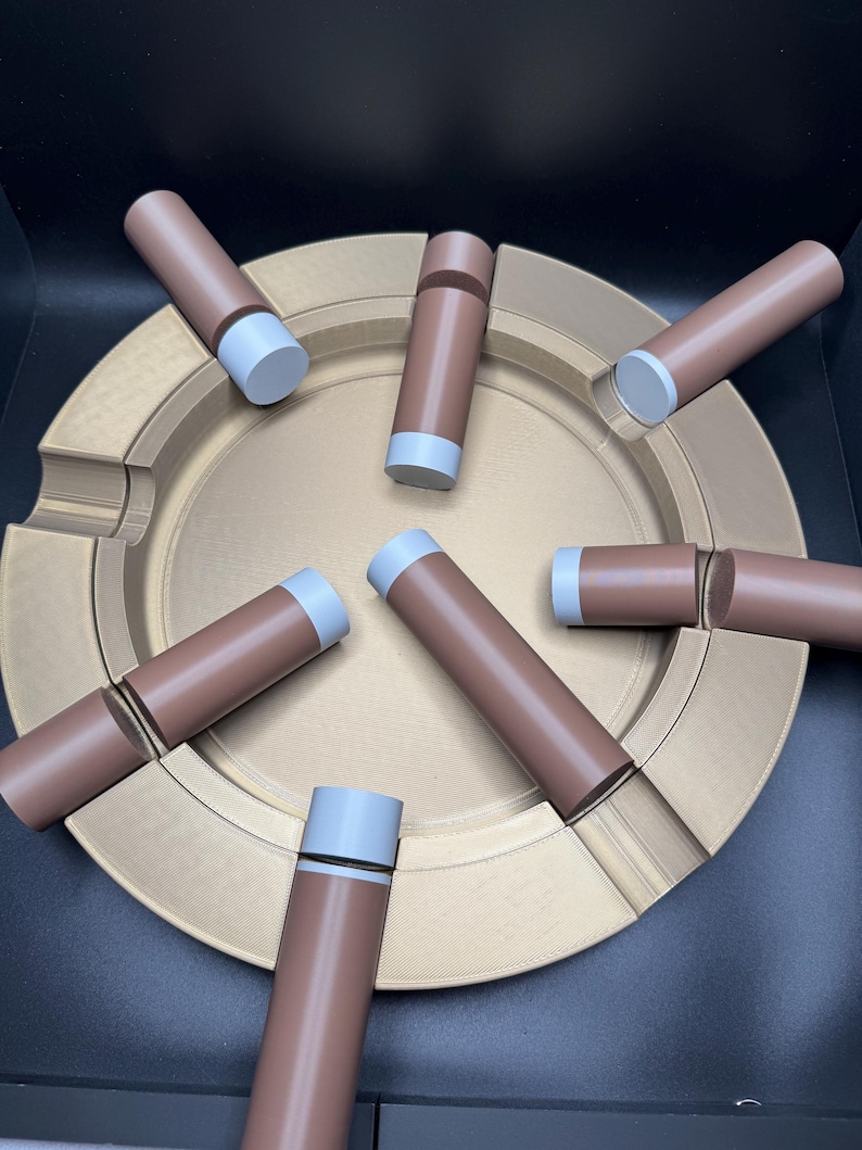 K&ouml;nnte beinhalten: Ein goldfarbener Aschenbecher mit kreisf&ouml;rmigem Design, der mehrere braune und graue zylindrische Objekte aufweist. Die Objekte sind um den Aschenbecher angeordnet. Der Aschenbecher hat eine strukturierte Oberfl&auml;che.