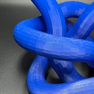 May include: A blue 3D printed knot sculpture. The knot is made of multiple intertwined loops, creating a complex and intricate design.