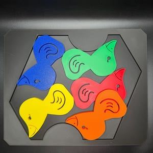 May include: A colorful puzzle featuring six bird-shaped pieces in blue, green, red, orange, and yellow. Each piece has a stylized ear design and a small hole. The pieces fit into a black hexagonal tray.
