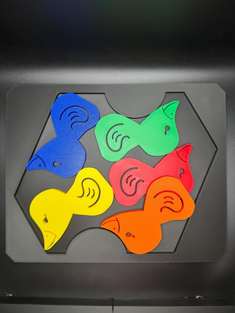 May include: A colorful puzzle featuring six bird-shaped pieces in blue, green, red, orange, and yellow. Each piece has a stylized ear design and a small hole. The pieces fit into a black hexagonal tray.