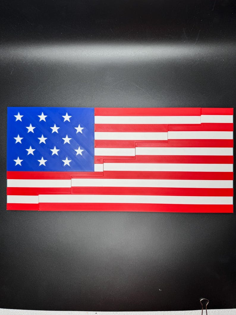 Puede incluir: Una bandera estadounidense impresa en 3D hecha de pl&aacute;stico rojo, blanco y azul. La bandera est&aacute; compuesta por piezas individuales que encajan para formar la bandera completa.