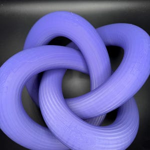 May include: A 3D-printed, interlocking purple torus knot. The knot is a continuous loop, with three interlocking rings. The object is made of a solid, textured material and is displayed against a dark background.