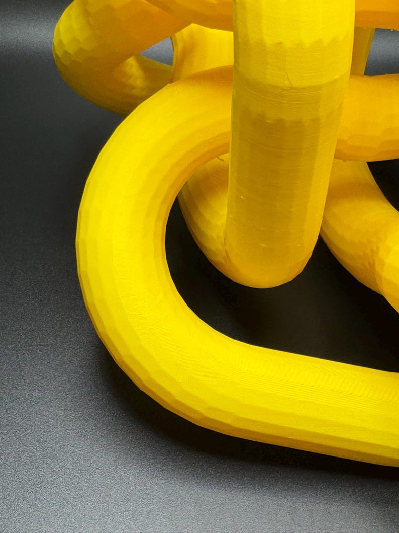 May include: A yellow 3D printed object with a complex, interwoven design. The object is made of plastic and has a smooth, glossy finish.