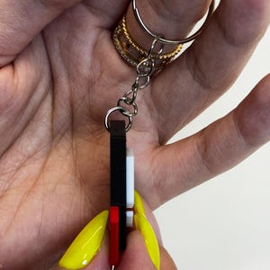 May include: A rectangular keychain with a black, white, and red design. The keychain is attached to a silver keyring. The image is taken from a close-up angle.