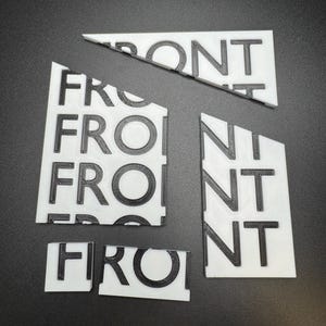 May include: White 3D-printed puzzle pieces spelling the word "FRONT" in black, with a textured surface. The pieces are arranged on a dark surface, with some pieces broken apart.