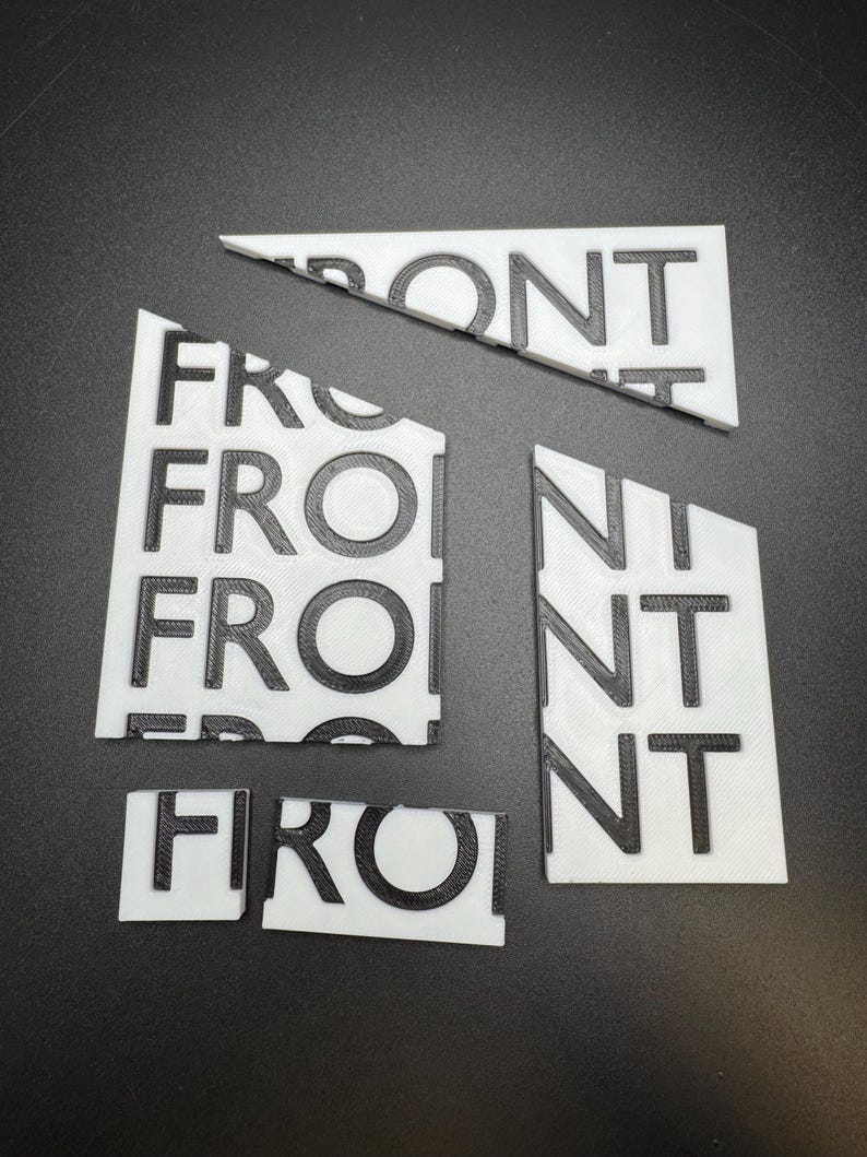 May include: White 3D-printed puzzle pieces spelling the word "FRONT" in black, with a textured surface. The pieces are arranged on a dark surface, with some pieces broken apart.