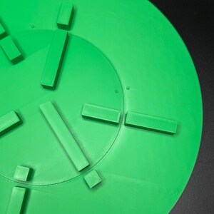 May include: A bright green, circular object with raised rectangular and square shapes arranged around the center. The object appears to be 3D-printed, with visible layer lines. The background is a solid black.