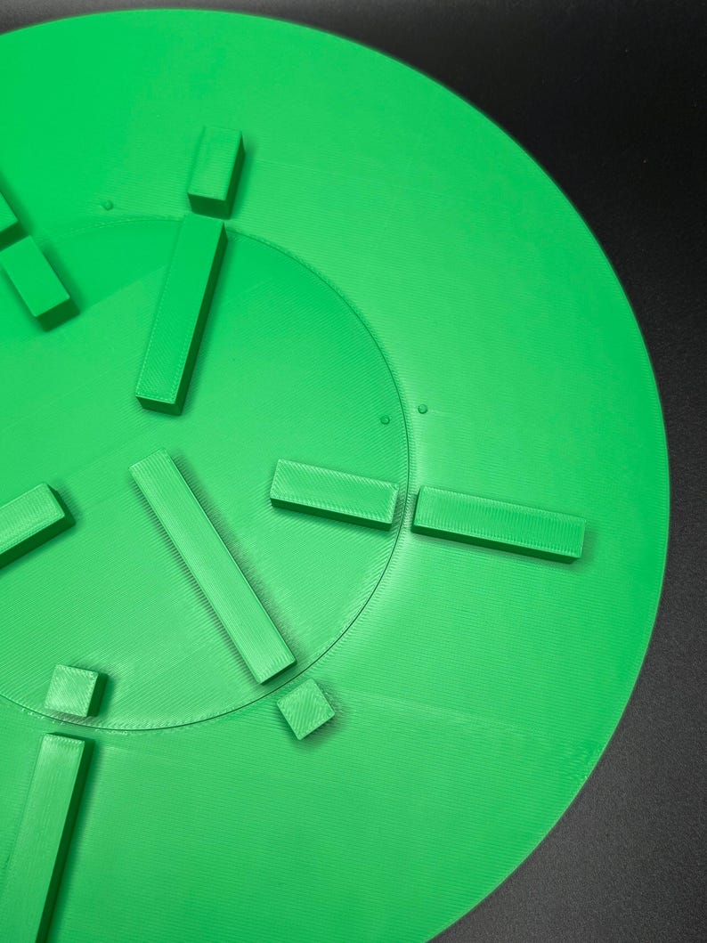 May include: A bright green, circular object with raised rectangular and square shapes arranged around the center. The object appears to be 3D-printed, with visible layer lines. The background is a solid black.