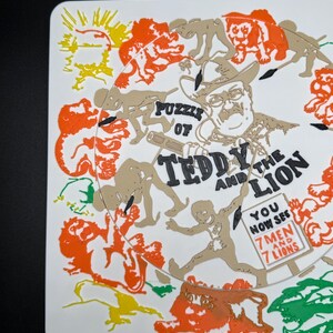 May include: A white square coaster with a colorful illustration. The design features a portrait of a man in a cowboy hat, surrounded by figures and lions. Text reads "Puzzle of Teddy and the Lion" and "You now see 7 men and 7 lions."