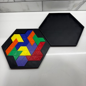Op de afbeelding: Een zwarte zeshoekige puzzeldoos met een deksel. De doos is gevuld met kleurrijke driehoekige puzzelstukjes in rood, oranje, geel, groen, blauw en paars.